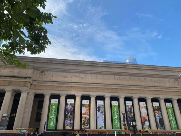 Front façade of Union Station with tall columns and green banners, blue sky and tree branches on the left.