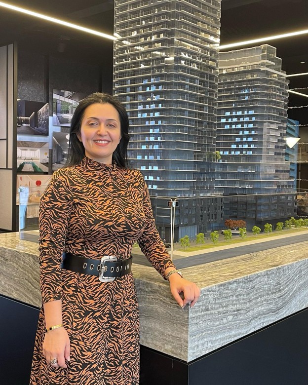 Picture10 - The Canada Times Woman in a leopard-print dress stands beside a detailed architectural scale model of modern skyscrapers, smiling at the camera.