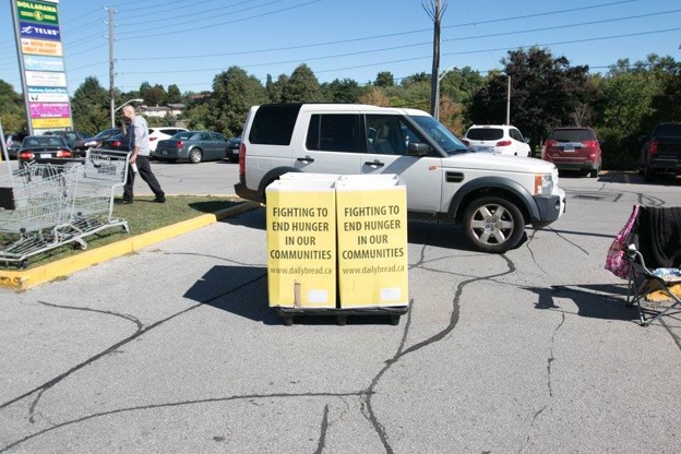 Two yellow donation boxes reading 'Fighting to End Hunger in Our Communities' on a cart in a parking lot, with a white SUV and shoppers nearby.