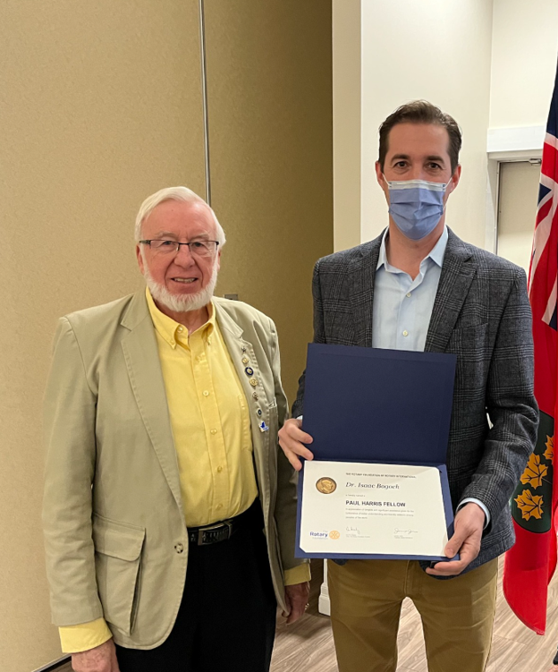 Picture13 - The Canada Times Two men pose for a photo as one, wearing a mask, holds an award certificate in a blue folder alongside an older man in a beige jacket and yellow shirt.