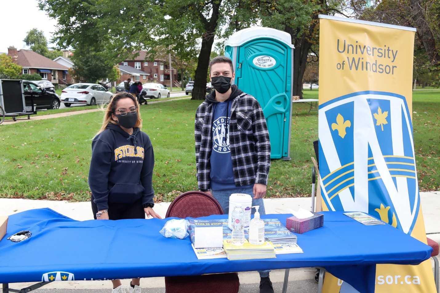 Picture17 - The Canada Times Two masked volunteers stand behind a blue campus information table at an outdoor University of Windsor event, with brochures and hand sanitizer on the table.