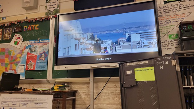 Picture22 - The Canada Times TV in a classroom displaying a movie scene: two people on a rooftop with a city and water in the background; subtitles read 'Shelby who?'