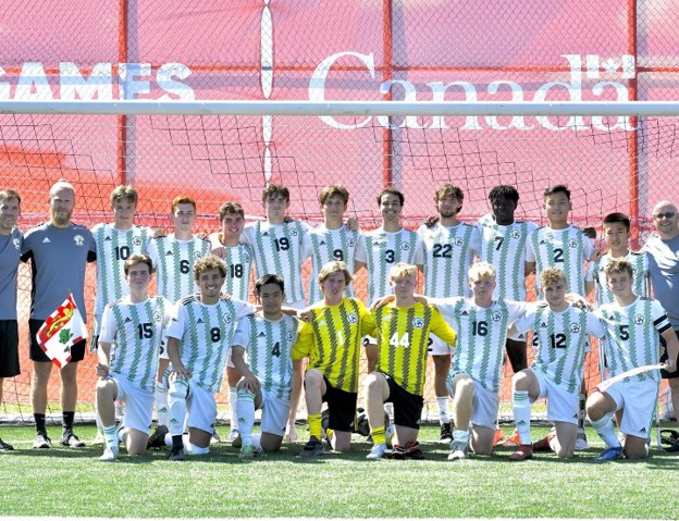 Group photo of a youth soccer team in white and green striped kits posing on a grass field, with two goalkeepers in yellow kneeling in front and coaches at the ends.