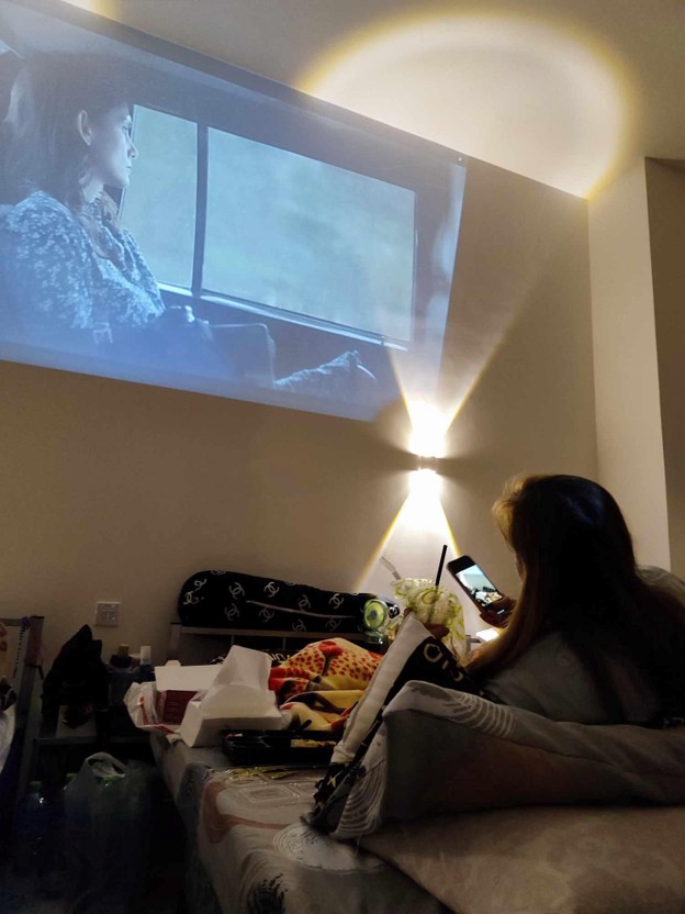 Picture19 - The Canada Times Woman lounging on a bed, watching a movie projected on the wall, while scrolling on her phone in a cluttered bedroom.