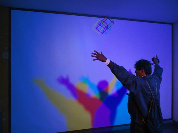 Picture31 - The Canada Times Person in a suit reaches toward a white wall where colorful shadows are projected by a hanging geometric sculpture, as part of a light art installation.