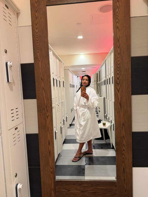 Picture32 - The Canada Times Woman in a white robe takes a mirror selfie in a locker room with rows of lockers behind her and checkered floor.