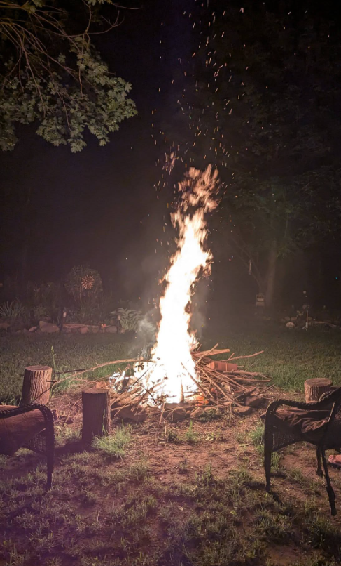 Picture33 - The Canada Times Nighttime outdoor scene with a tall bonfire surrounded by wooden logs and chairs on grass.