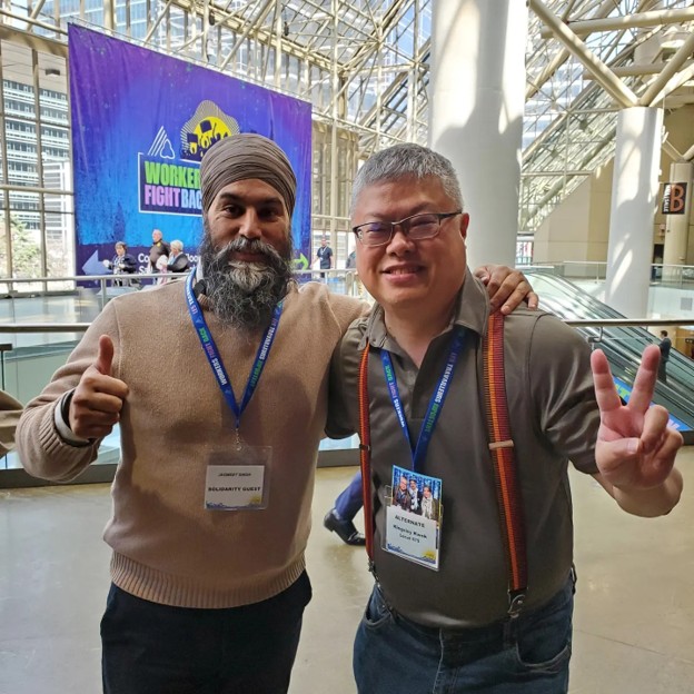 Picture6 - The Canada Times Two men at a conference pose for a photo, one with a turban and beard, both wearing badges, giving thumbs up and a peace sign in front of a ‘Workers Fight Back’ banner.