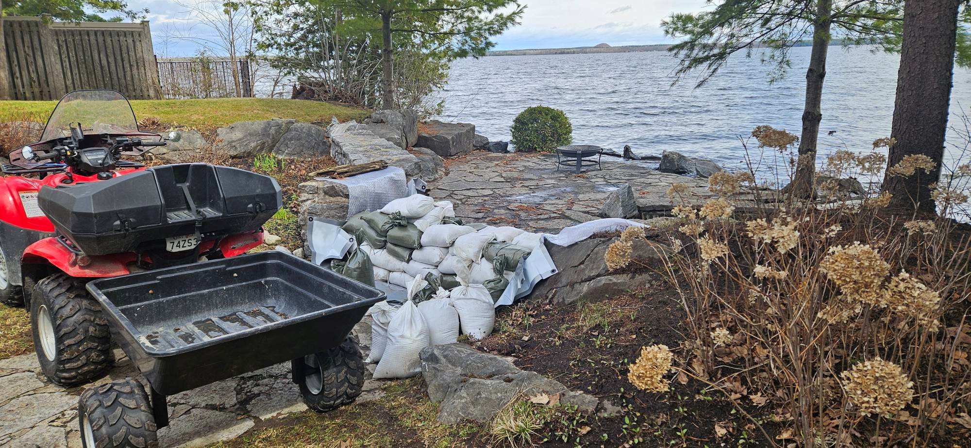 Red ATV with an empty hauling tub parked on a stone patio by a lake, with sandbags and rocks nearby.