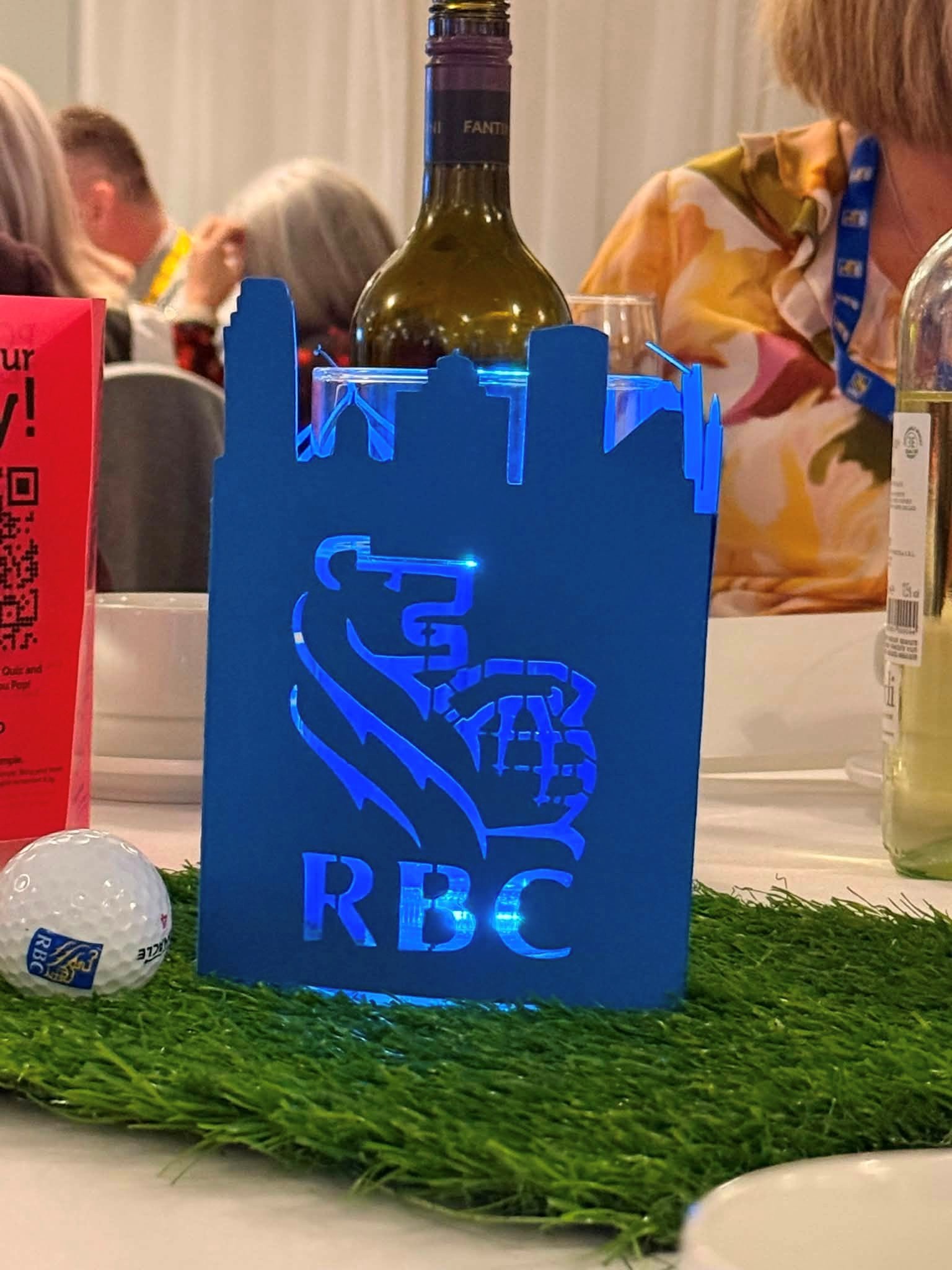 10 - The Canada Times Blue illuminated RBC centerpiece with skyline cutout and hand logo on a green mat beside a wine bottle.