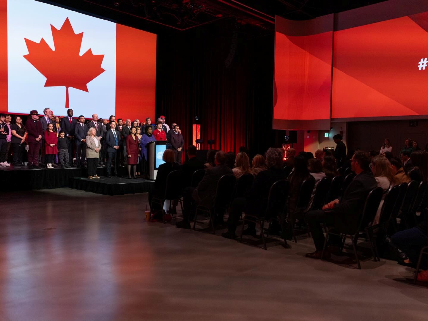 13 - The Canada Times Group of people standing on a stage in front of a large Canadian flag during a ceremony, audience seated in the darkened auditorium.