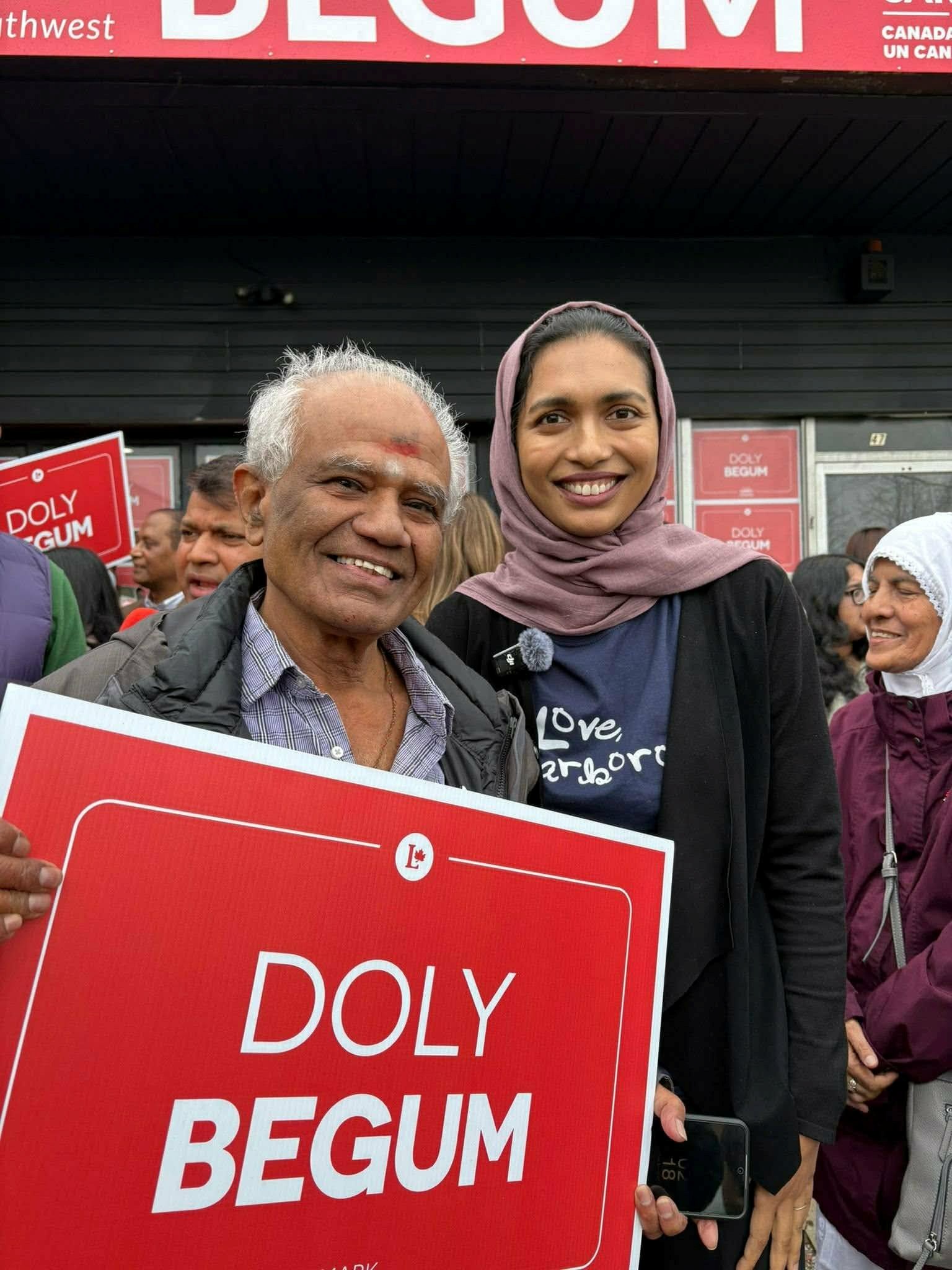 Smiling man and woman in a hijab holding a red poster that reads ‘DOLY BEGUM’ at a public rally.