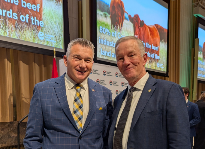 Two men in business suits posing for a photo at a conference, with large screens showing cattle in the background.