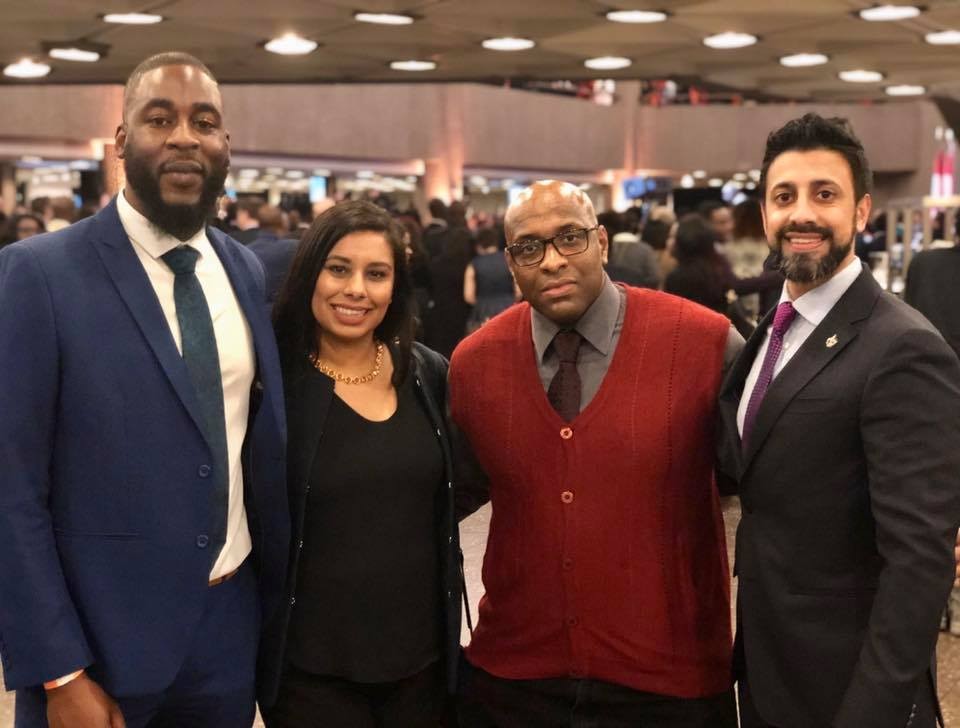 5 - The Canada Times Four professionals posing together at a busy indoor event, smiling at the camera (two men in suits, a woman in black, and a man in a red sweater vest).
