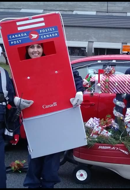 5 - The Canada Times Person wearing a red Canada Post mailbox costume with their face visible, standing next to a decorated red car at a parade.