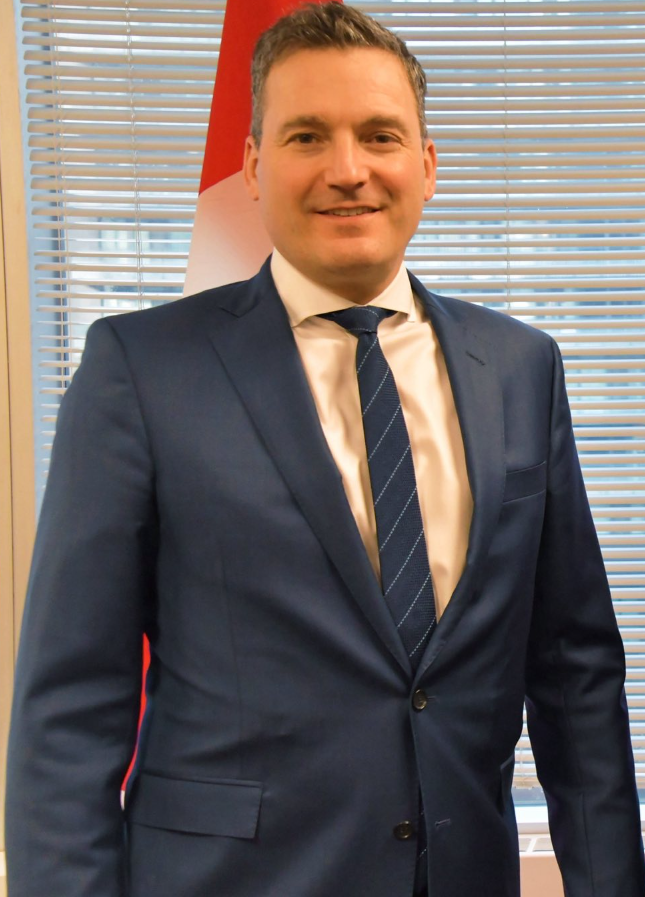 6 - The Canada Times Professional portrait of a man in a navy suit and tie, standing in front of blinds and a red-and-white flag behind him.