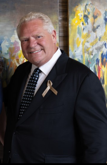Smiling older man in a dark suit and patterned tie, wearing a ribbon pin, standing against colorful abstract paintings.