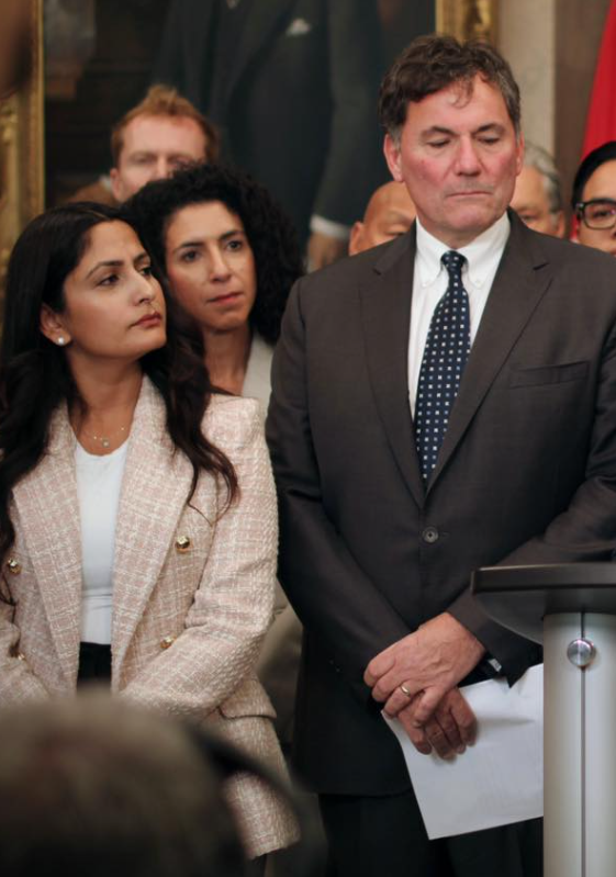 A woman in a pale blazer and a man in a dark suit stand near a podium at a formal event with others in the background.