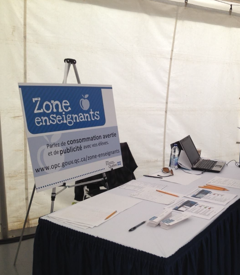 7 - The Canada Times Blue and white sign 'Zone enseignants' on a stand beside a table with papers, pens, a laptop and a water bottle under a white tent.