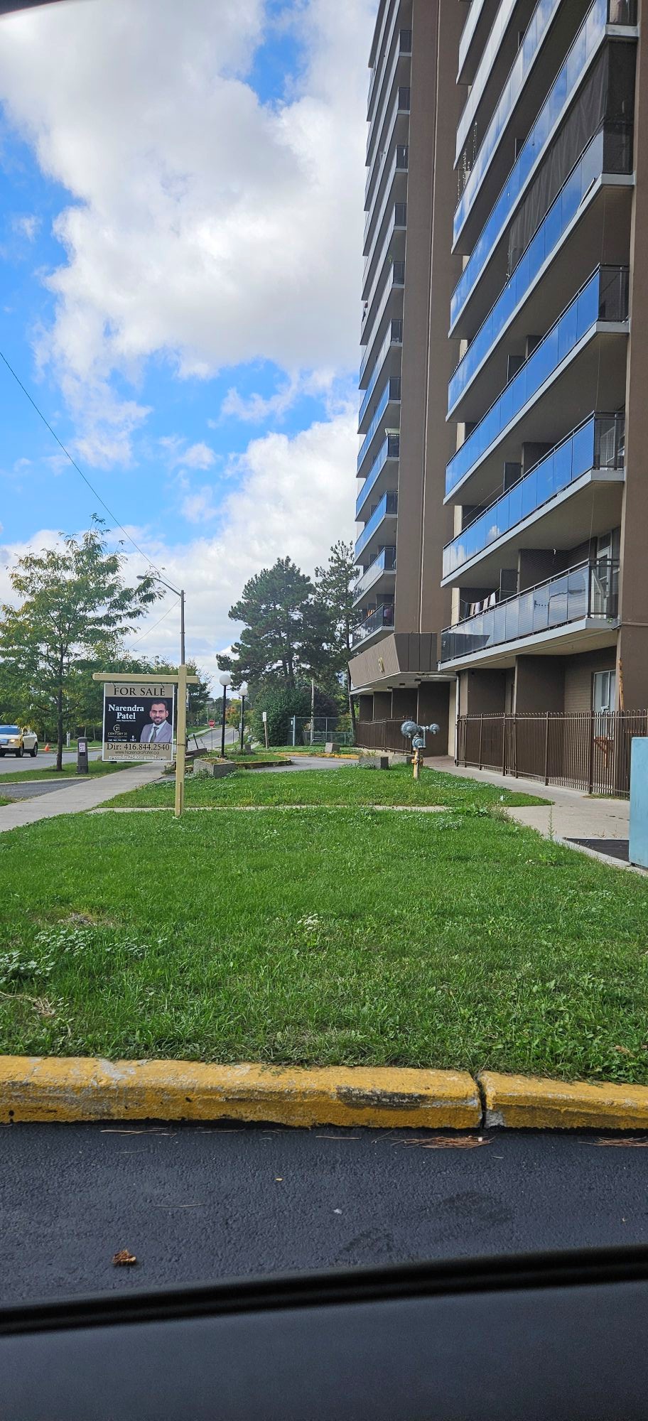 High-rise brown apartment building with blue-glass balconies on the right, a grassy foreground, and a 'For Sale' sign featuring a realtor's photo and the name Narendra Patel nearby.
