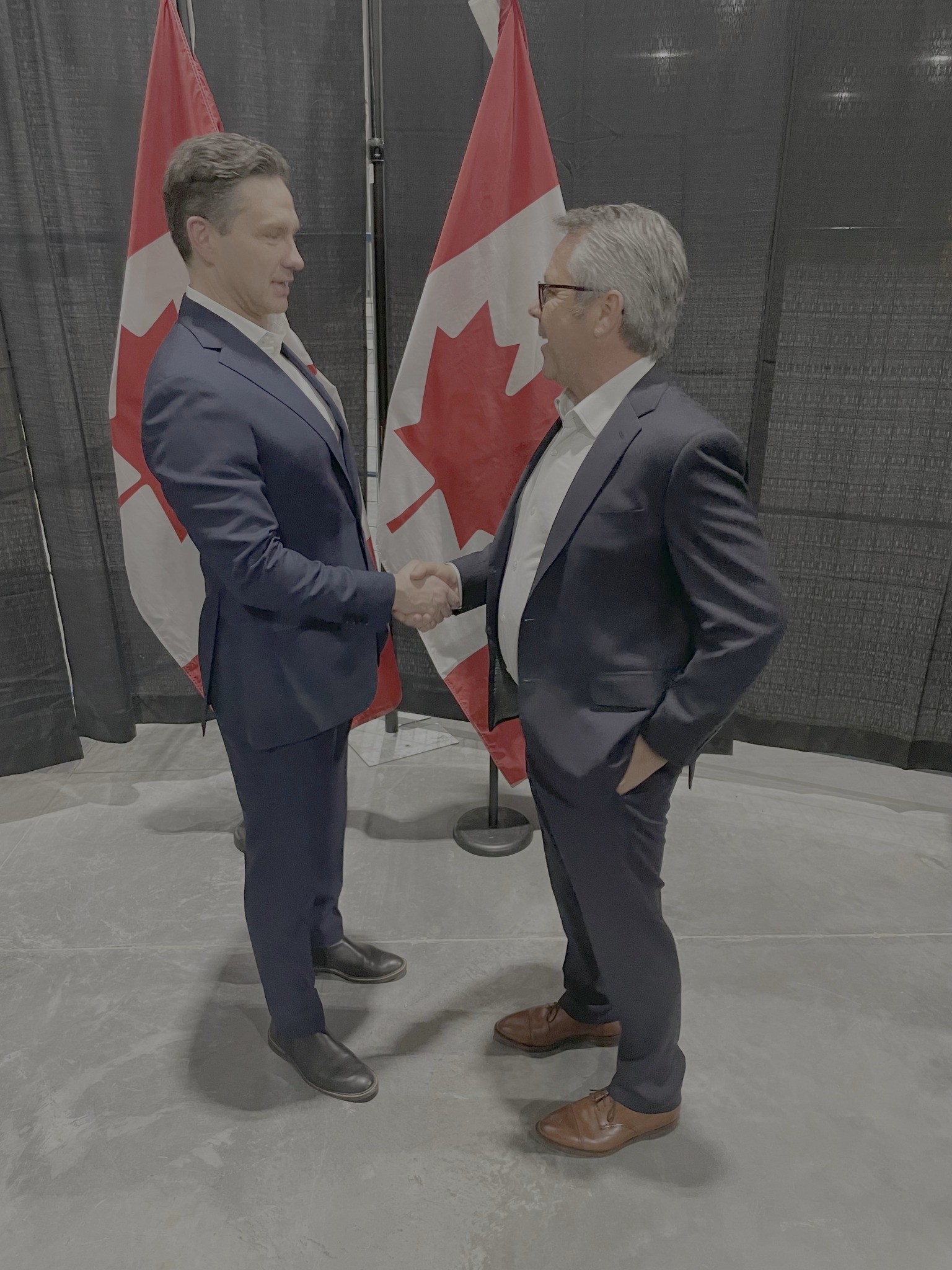 Two men in suits shake hands in front of Canadian flags at a formal event.