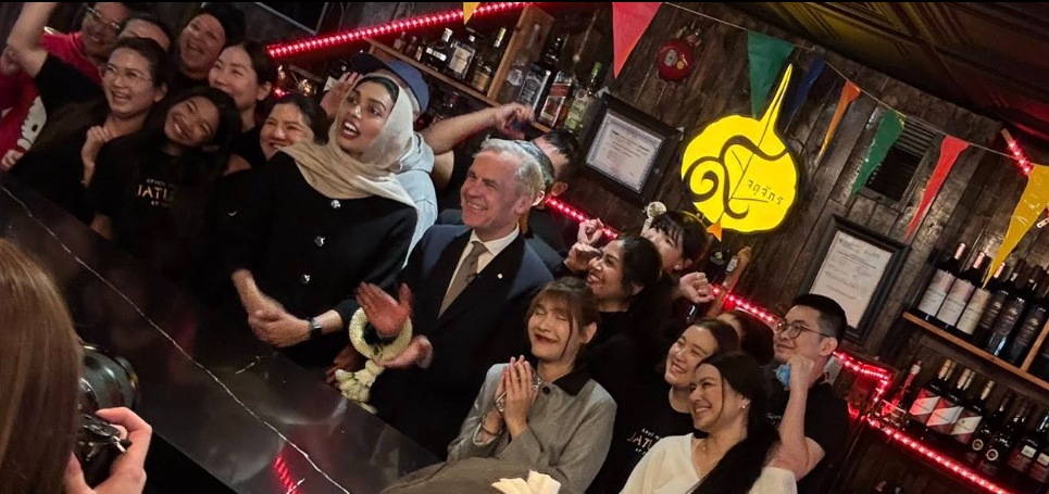 9 - The Canada Times Group of smiling people posing together at a bar or event counter with colorful lights and flags in the background, some clapping or posing for a photo.
