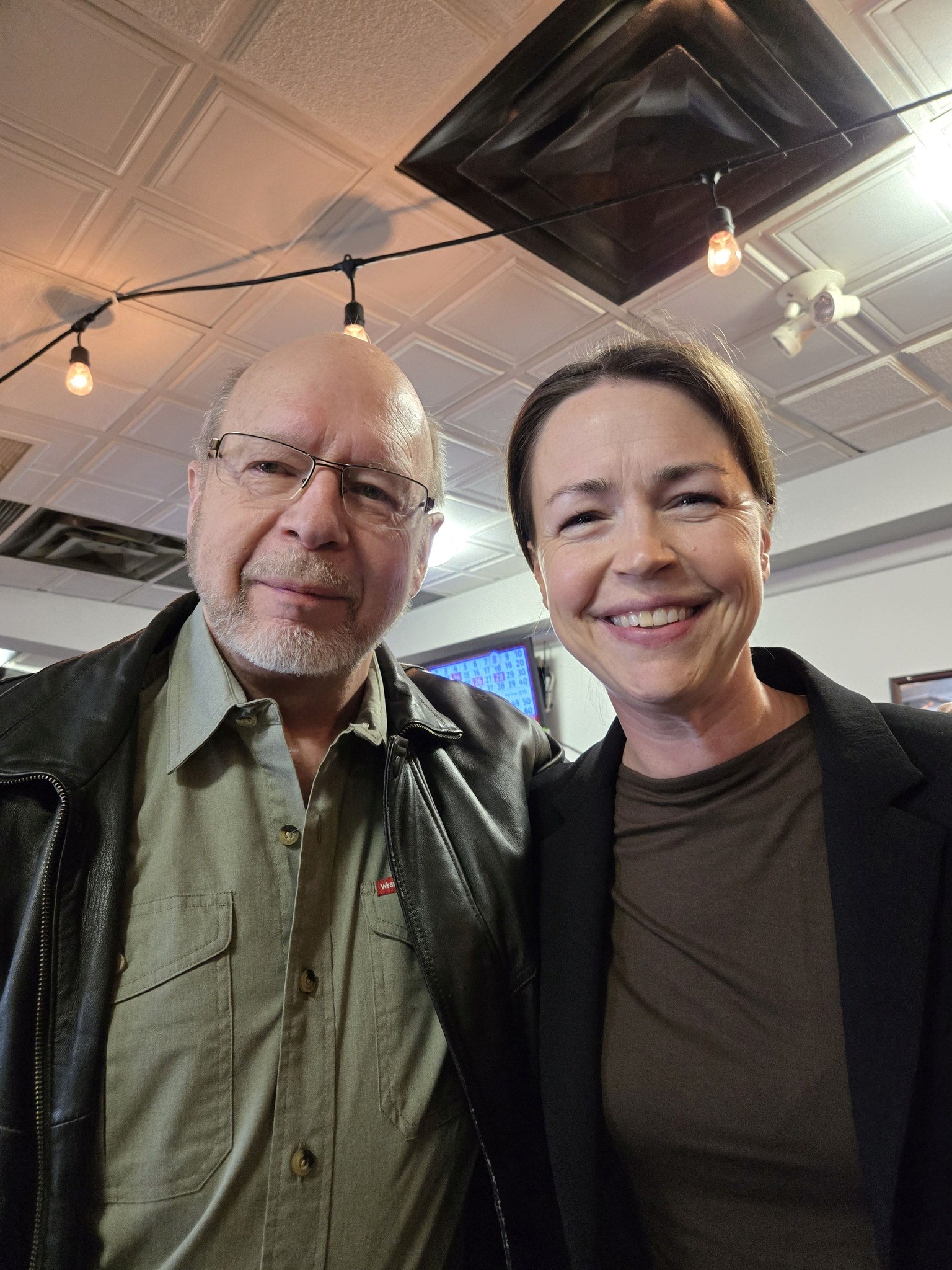 1 - The Canada Times Two smiling adults taking a selfie indoors, man with glasses on the left and woman on the right, ceiling with string lights above them.