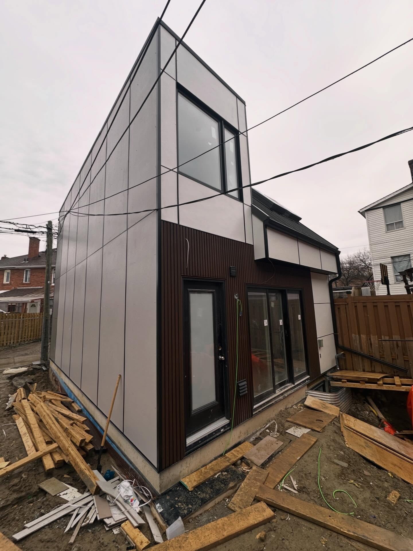 Modern two-story extension with gray metal panel siding and a large corner window, under construction with wood planks and debris on the ground.