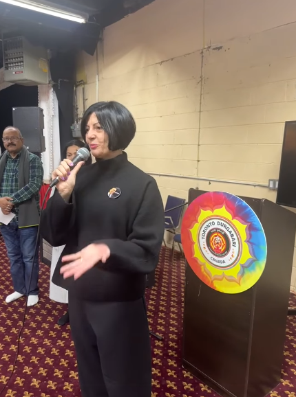 10 - The Canada Times Woman with a short black bob speaks into a microphone at an indoor event, standing near a podium with a colorful circle emblem behind her.
