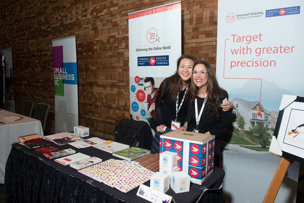 11 - The Canada Times Two smiling women stand behind a booth at a trade show, with banners reading 'Target with greater precision' and 'Delivering the Online World' nearby.