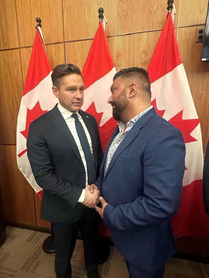 13 - The Canada Times Two men in suits shake hands in front of Canadian flags in a formal setting, facing each other and smiling slightly.