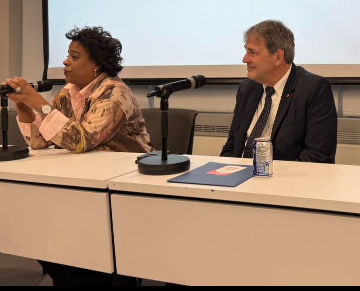 13 - The Canada Times Two panelists sit at a white table with microphones, the woman speaking on the left and the man in a suit listening on the right in a conference room.