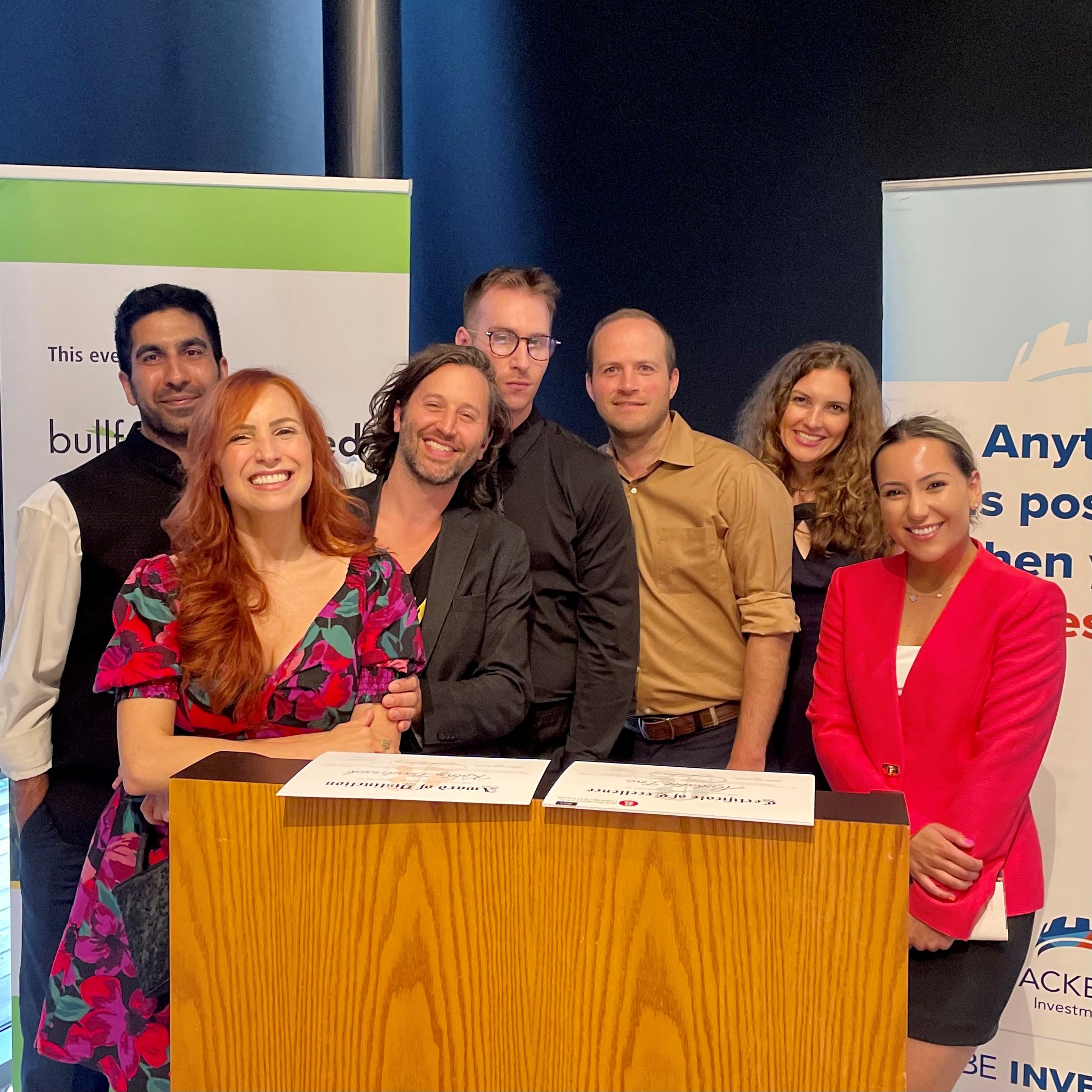 2 - The Canada Times Group of smiling people behind a wooden desk at a booth during an event, with banners in the background.