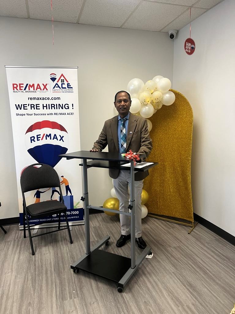 Man in a brown suit stands at a black mobile podium in a real estate office, with a RE/MAX Ace banner saying "WE'RE HIRING!" and balloons in the background.
