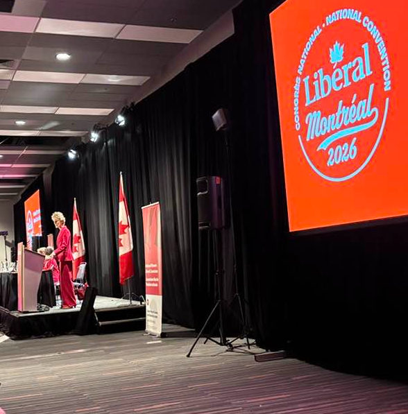 Female speaker at a podium on stage during a Liberal event, with red banners, Canadian flags, and a large red 'Liberal Montréal 2016' screen.