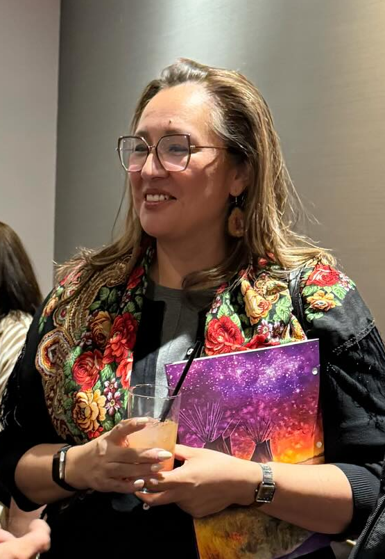 Woman with glasses and floral scarf smiles while holding a purple booklet and a glass of orange juice at a social event.