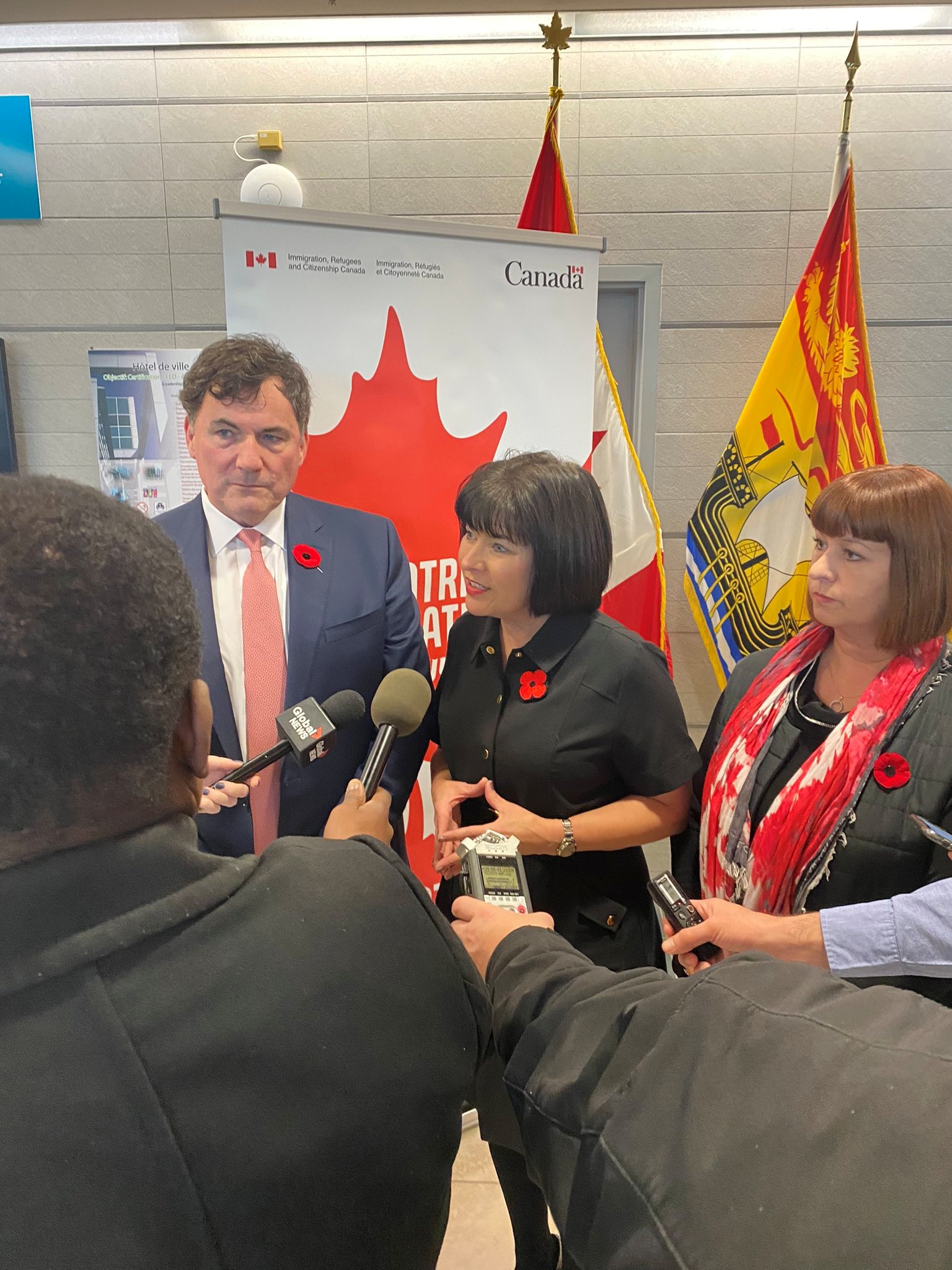 6 - The Canada Times Politician in a navy suit and pink tie speaks to reporters, Canada flag and banner behind him
