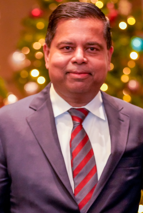 Portrait of a man in a purple suit and red striped tie, smiling with blurred Christmas lights in the background at a holiday event.