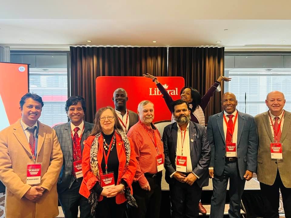 7 - The Canada Times Group of diverse professionals posing for a photo at a conference, red Liberal backdrop behind them and name badges visible on lanyards.