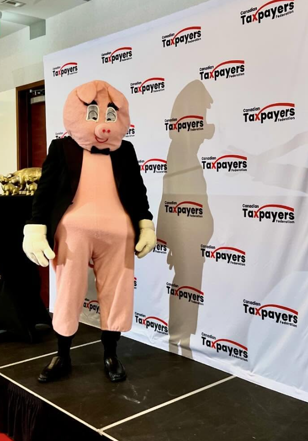 7 - The Canada Times Pig mascot in a pink suit and black jacket stands on a stage beside a Taxpayers Federation backdrop.