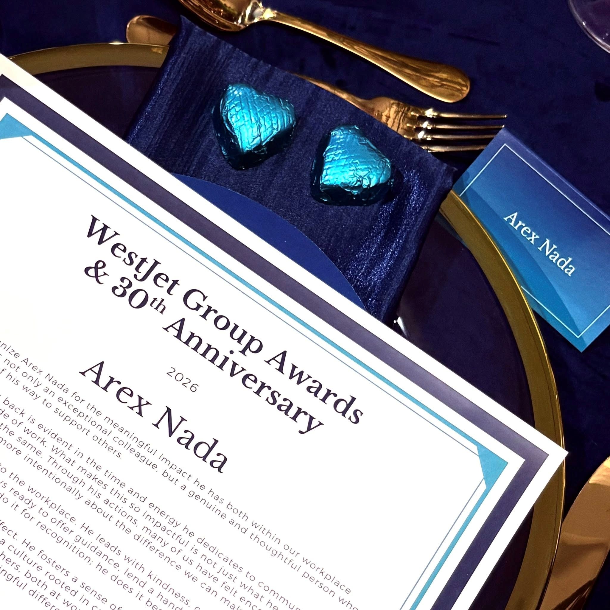 Close-up of an award invitation with teal foil hearts, blue-bordered card and gold cutlery on a dark blue fabric background.