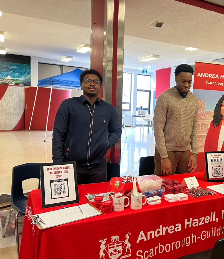 8 - The Canada Times Two men stand behind a red information table for Andrea Hazell Scarborough-Guildward event, with volunteer sign-up materials and QR codes on display.