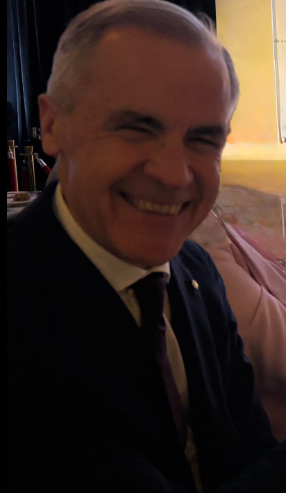 9 - The Canada Times Smiling older man in a dark suit and tie, seated indoors, with a warm yellow background behind him.