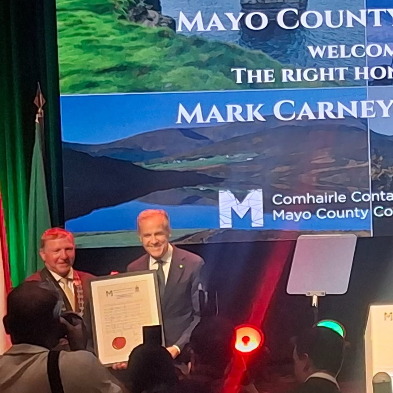 1 - The Canada Times Two men in suits on a stage presenting a framed certificate at an award ceremony, with a Mayo County backdrop in the background.