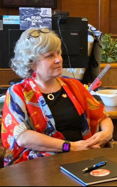 1 - The Canada Times Older woman with short gray hair, glasses on her head, wearing a colorful red‑and‑blue shawl over a black top, seated at a table in a wood-paneled room.