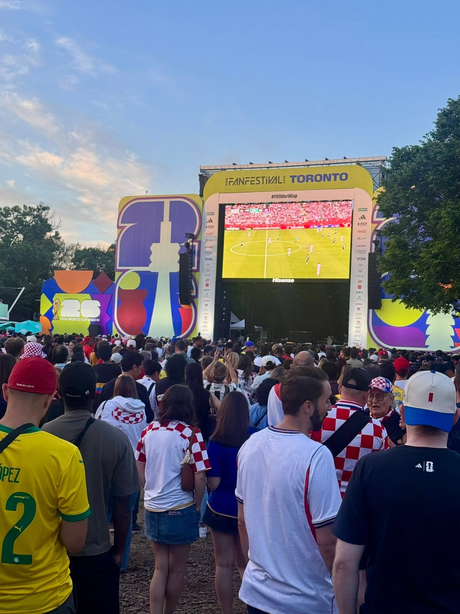 Large outdoor crowd watching a soccer match on a giant screen at a FIFA World Cup festival in Toronto, with colorful festival decor nearby and clear blue sky.