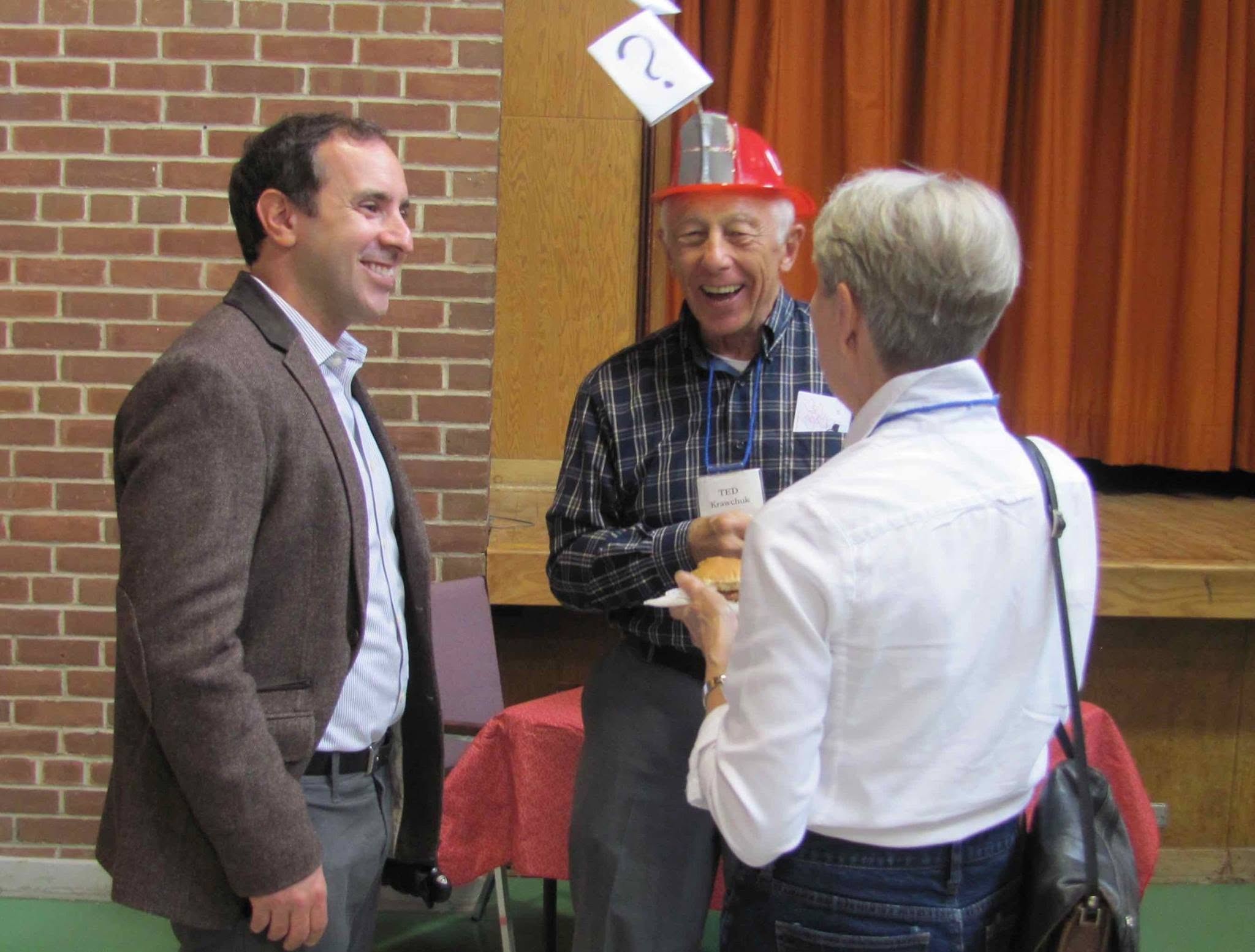 1 - The Canada Times Three adults standing and chatting indoors: a smiling man in a brown blazer on the left, a man in a plaid shirt with a red hard hat in the middle, and a woman with short gray hair facing away on the right.