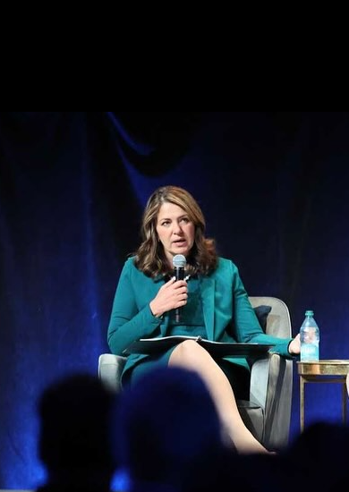 1 - The Canada Times Woman in a teal blazer sits on stage speaking into a handheld microphone during a panel discussion, with a water bottle on a side table nearby.