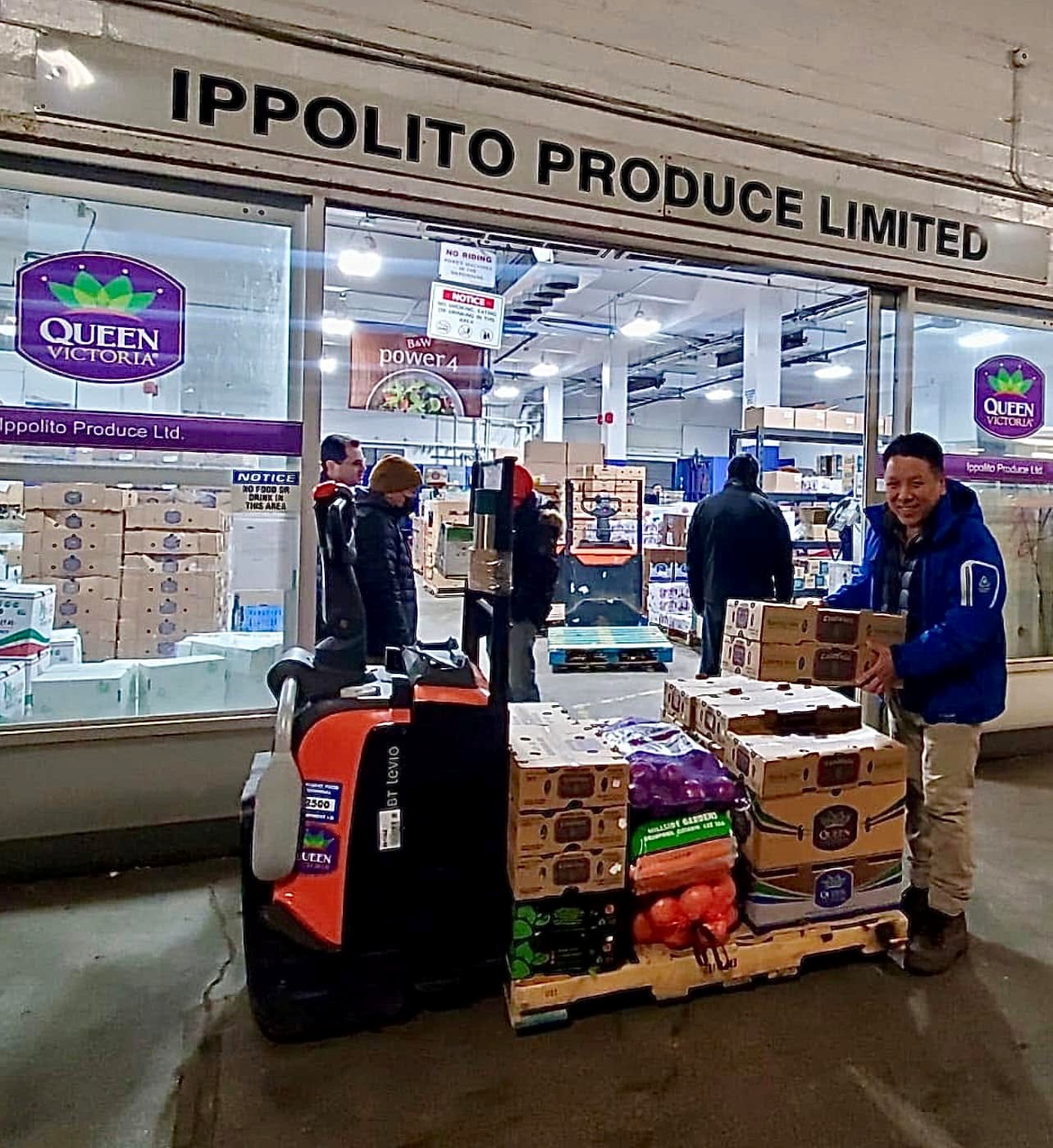 10 - The Canada Times Man in a blue jacket hands full, lifting boxes onto a pallet in front of Ippolito Produce Limited storefront.