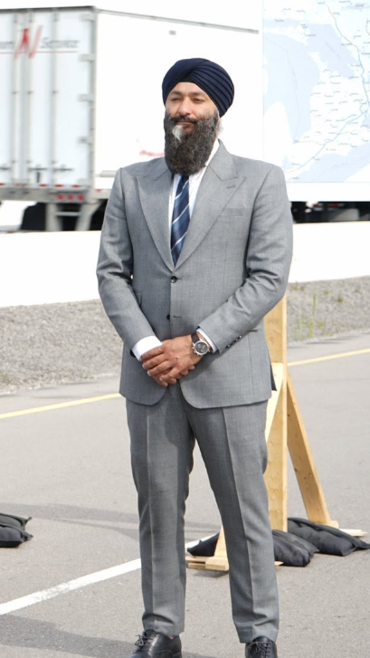 Man in a grey suit and navy turban stands with hands clasped on a road, trucks and a map in the background.